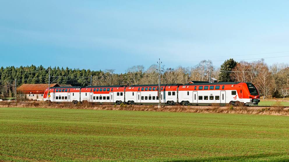 Tågmodellen ER1 ute på spåret (Tåg i Bergslagen). Dubbeldäckartåg med vit och röd målning åker genom landskap med grön åker i förgrunden och lätt blå himmel bakom.