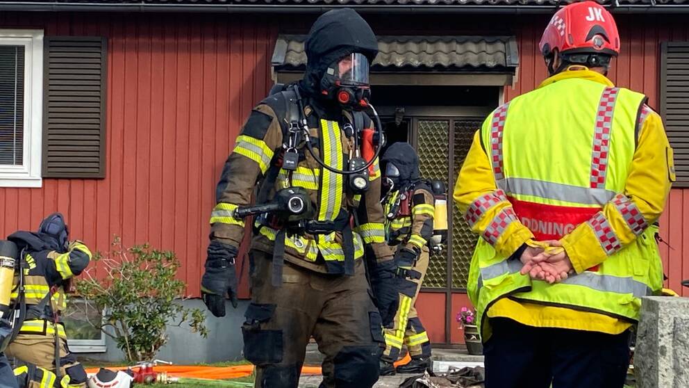 Rökdykare med svart huva och mask för ansiktet framför rött hus. Räddningsledare i gul jacka med röd hjälm står med ryggen mot kameran. Jämjö, Trädgårdsvägen, villabrand, övertänd.
