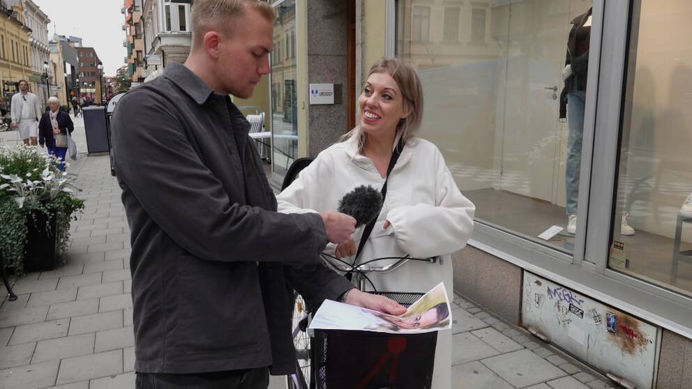 En man står i stadsmiljö iförd en grön skjortjacka. Han håller i en mikrofon och i en bild på en person som han visar upp för en blond kvinna i vit tröja som går med en cykel. Kvinnan skrattar lite lätt och tittar på mannen.