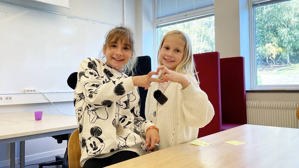 två 9-åriga flickor i vita tröjor, inne i ett klassrum håller fingrar mot varandra som bildar ett hjärta