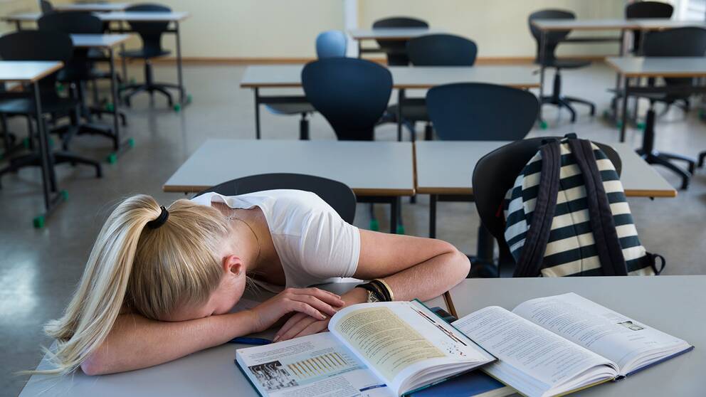 Tonårstjej ligger över bänken i klassrum. Framför henne ligger böcker.