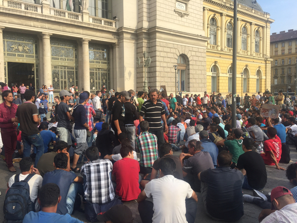 Just nu utanför Keleti, människor sitter i väntan på ny information om vad som ska ske härnäst. En ungersk man talar i megafon om hur stolt han är över att vara här tillsammans med de som väntar.