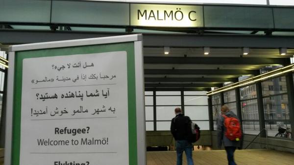 Hej jag är vid Malmö centralstation och den här och liknande skyltar välkomnar flyktingarna till Malmö. I närheten finns en samlingsplats där Röda korsets personal och andra frivilliga hjälper till med mat och kläder.