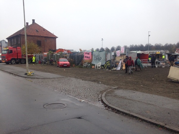 Det råder en låst och märklig situation vid lägret nu. De boende sitter kvar vid ingången. Samtidigt rullas den stora rivningsmaskinen in genom stängslet strax intill.